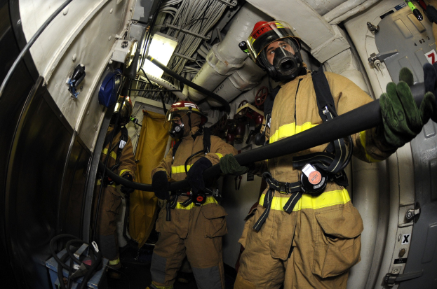 Feuerwehrleute in Helmen und Handschuhen mit einer Schlauchleitung in einem Flugzeug mit Ausrüstung und Text im Hintergrund.