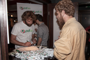 Eine Gruppe von Menschen, die um einen Tisch mit Ausweisen und Papieren stehen, einer hölt einen Stift, mit einer offenen Tür und einem Banner im Hintergrund.