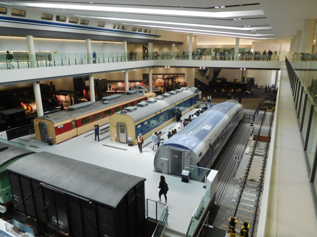 Ein geräumiges Museumsausstellungsstück, das verschiedene Züge in verschiedenen Größen und Farben zeigt, mit Besuchern, die vom Boden, Geländern und Säulen unter Deckenlampen beobachten.