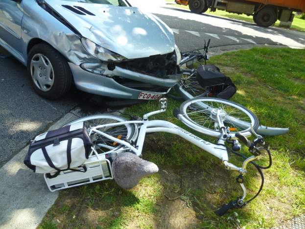 Ein verunglücktes Auto am Straßenrand mit einem Fahrrad auf dem Gras, Bäume und einen klaren blauen Himmel im Hintergrund.