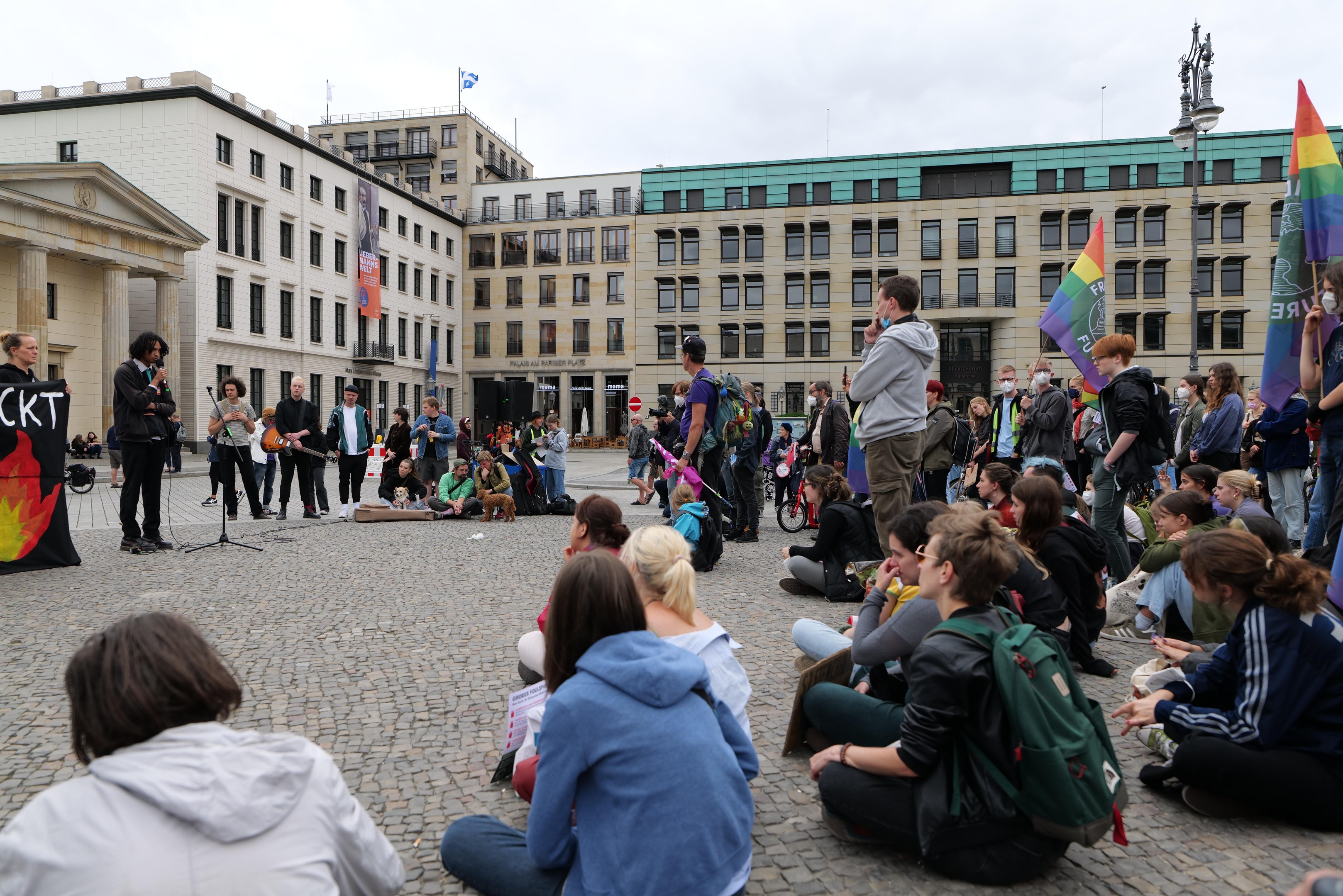 Eine Gruppe von Menschen, die auf dem Boden sitzen, vor einer Menge mit Fahnen und Spruchbändern während einer anti-schwulen Demonstration in Berlin, mit einer Statue, Gebäuden und einem Mikrofonständer im Hintergrund.