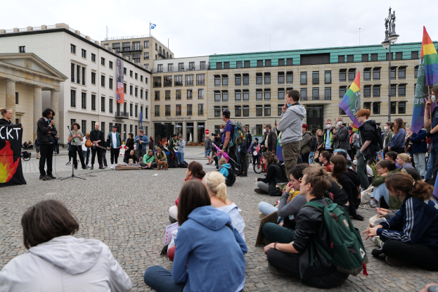 Eine Gruppe von Menschen, die auf dem Boden sitzen, vor einer Menge mit Fahnen und Spruchbändern während einer anti-schwulen Demonstration in Berlin, mit einer Statue, Gebäuden und einem Mikrofonständer im Hintergrund.