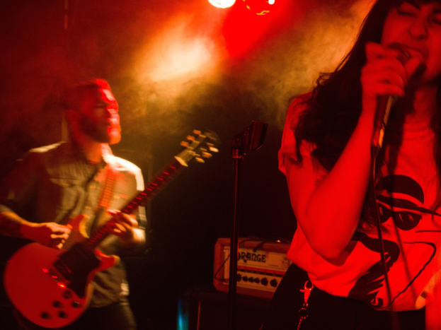 Eine Frau singt in ein Mikrofon auf der Bühne, wöhrend ein Mann eine Akustikgitarre im Hintergrund spielt, mit Deckenleuchten, die die Szenerie erhellen.