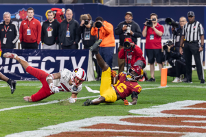 Ein Fußballspieler taucht in die Endzone für einen Touchdown ein, mit einer Gruppe von Menschen am Boden, einige halten Kameras, und ein Banner mit Text im Vordergrund und Geländer, Pfosten und andere Gegenstände im Hintergrund.