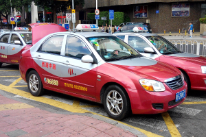 Zwei rote Taxis nebeneinander auf einer Straße mit Passanten, einem Zaun, Schildern, Bäumen, Gebäuden und Wandplakaten.