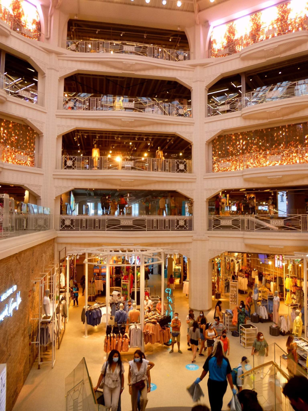 Großer Atrium eines belebten Einkaufszentrums mit Menschen, die gehen, Taschen tragen und Schaufensterpuppen mit Kleidern ansehen, umgeben von Geländern, Lichtern und Texttafeln an den Wänden.