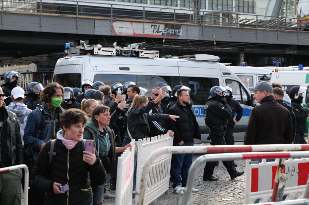 Gruppe von Menschen vor Polizeifahrzeugen mit Absperrungen im Vordergrund, Brücke und Gebäude im Hintergrund bei einer Protestauflösung in Berlin.