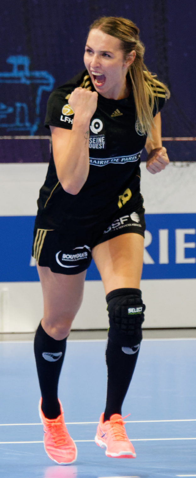 Eine Frau in schwarzem Shirt und Shorts, die Volleyball auf einem Court spielt, mit einem Banner im Hintergrund und Schuhen.