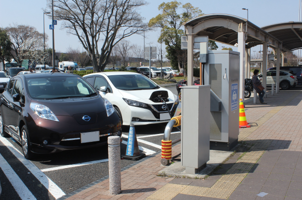Elektroauto-Ladestation in Japan mit Autos auf der Straße, Verkehrskegel, einer Person auf dem Gehweg und umgebender Infrastruktur vor einem Himmel als Hintergrund.