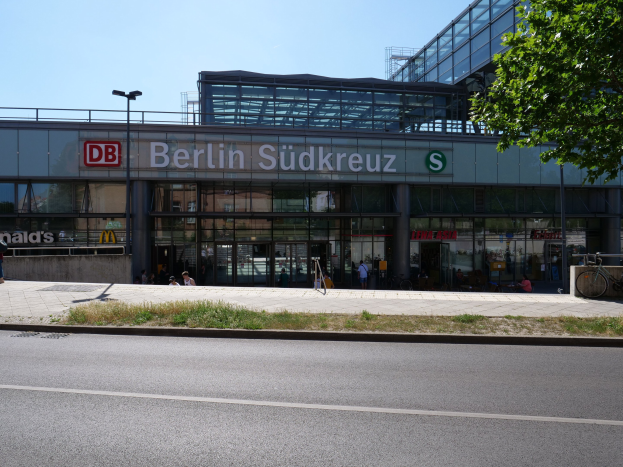 Großes gläsernes Berliner Südkreuz-Bahnhofsgebäude mit Text an seiner Fassade, umgeben von Straßenmasten, Laternen, Fahrzeugen, Fußgängern, Fahrrädern, Bäumen und einem klaren blauen Himmel.