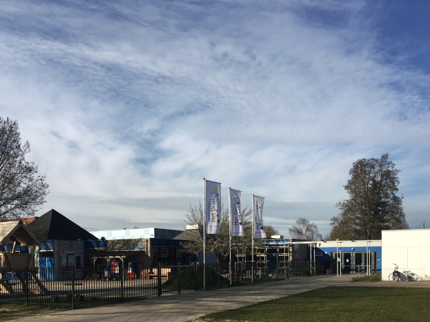 Großes Gebäude mit Metallzaun, Rasenfläche, Weg, Bäumen, Pfosten, Bannern, einem geparkten Fahrrad, nahen Häusern und einem bewölkten Himmel, identifiziert als der neue Spielbereich für Kinder an der Schule.