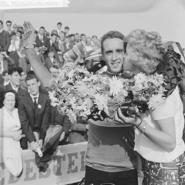 Ein Schwarz-Weiß-Foto eines Mannes und einer Frau, die sich in einer Menge umarmen und küssen, wobei der Mann einen Blumenstrauß hält und die Frau lacht, während die Menge applaudiert.