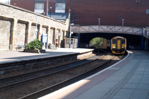 Ein Zug fährt auf Schienen mit Plattformen auf beiden Seiten, eine Hauswand links, Straßenlaternen, Pflanzen, Bänke, eine Kabine, eine Brücke über die Plattformen und Gebäude und Masten im Hintergrund.