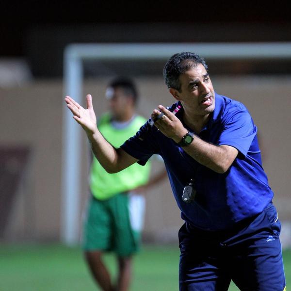 Ein Mann in einem blauen T-Shirt und Hose steht auf einem Fussballfeld und hält ein Mikrofon in der Hand, während er auf Spieler zeigt. Im Hintergrund ist eine Person in einem grünen Kleid, ein Torpfosten und eine Wand zu sehen.