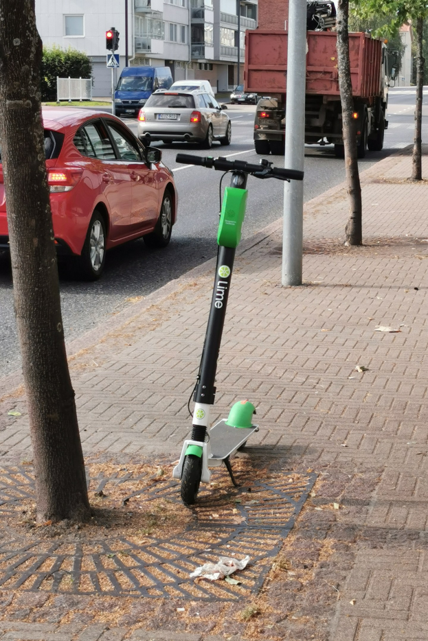Elektroroller auf einer Straße neben einem Baum geparkt, mit Fahrzeugen auf der Straße und Gebäuden im Hintergrund.