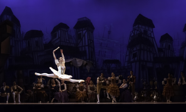 Eine Ballerina in einem weißen Tutu und Spitzenschuhen tanzt auf der Bühne, während ein Publikum zusieht, mit einem Burgenbild im Hintergrund.