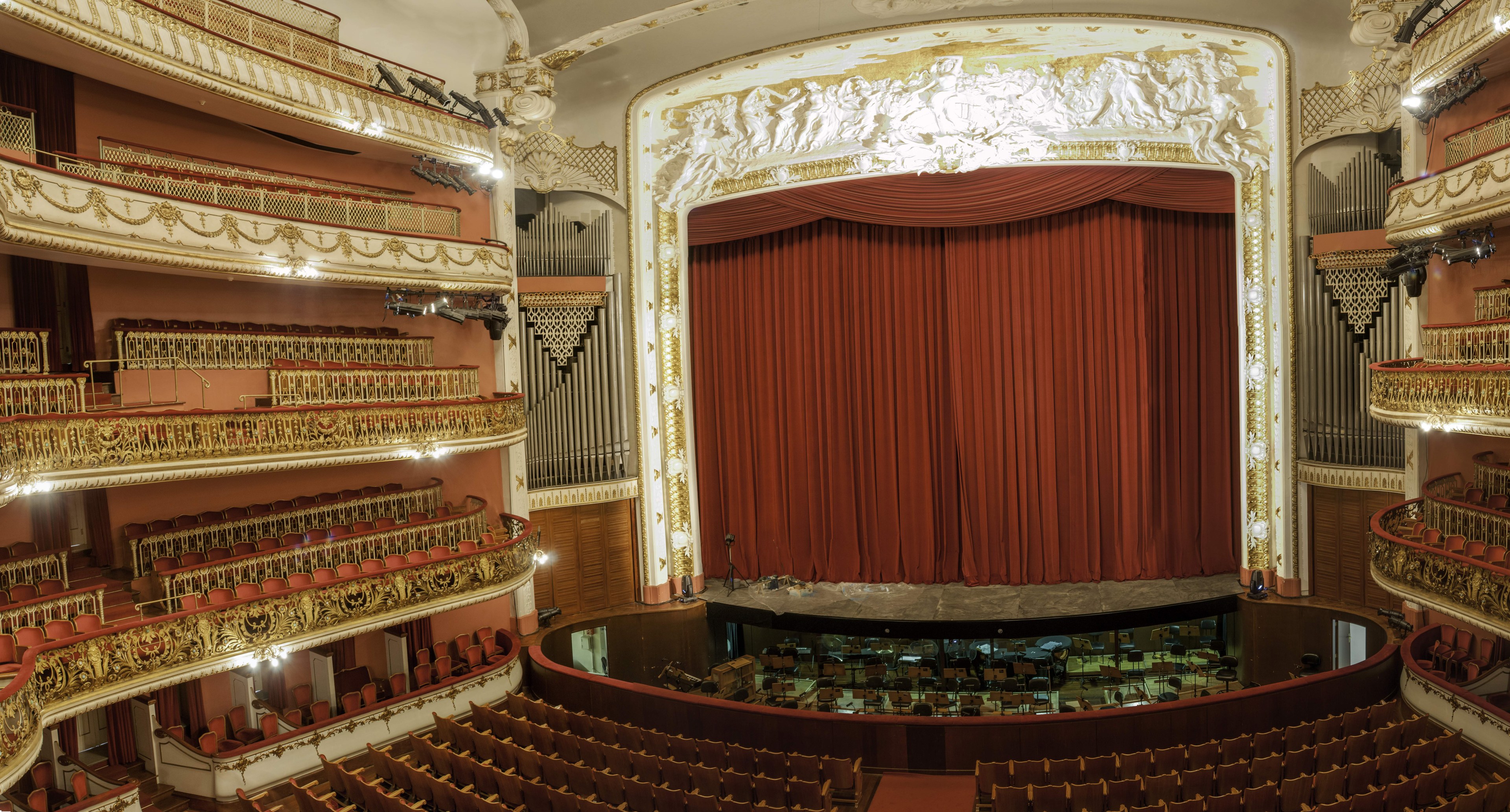 Großer Zuschauerraum mit roter Gardine, Stühlen, Geländern, Lampen und Wandskulpturen, mit Bühnenobjekten, die eine Konzerthalle vermuten lassen.