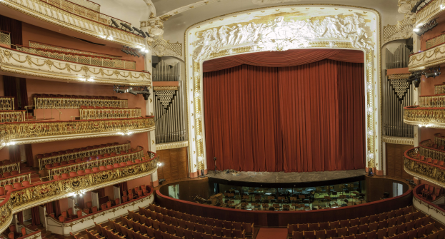 Großer Zuschauerraum mit roter Gardine, Stühlen, Geländern, Lampen und Wandskulpturen, mit Bühnenobjekten, die eine Konzerthalle vermuten lassen.