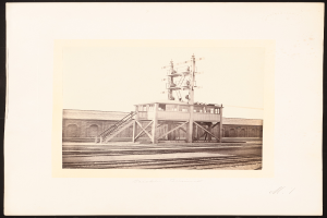 Ein Schwarz-Weiß-Foto eines Bahnhofs mit einer zentralen Signalbox, umgeben von Eisenbahnschienen und einem Gebäude im Hintergrund, mit Text unten.