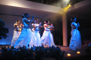 Eine Gruppe von Menschen tanzt auf einer Bühne mit Pflanzen davor und eine andere Gruppe von Menschen schaut zu.