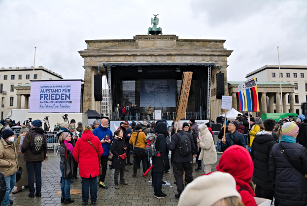 Eine Menschenmenge steht vor einem Gebäude mit einer Bühne, auf der Redner und ein Bildschirm zu sehen sind, umgeben von Flaggen und Transparenten mit Text, in Berlin während einer Demonstration.