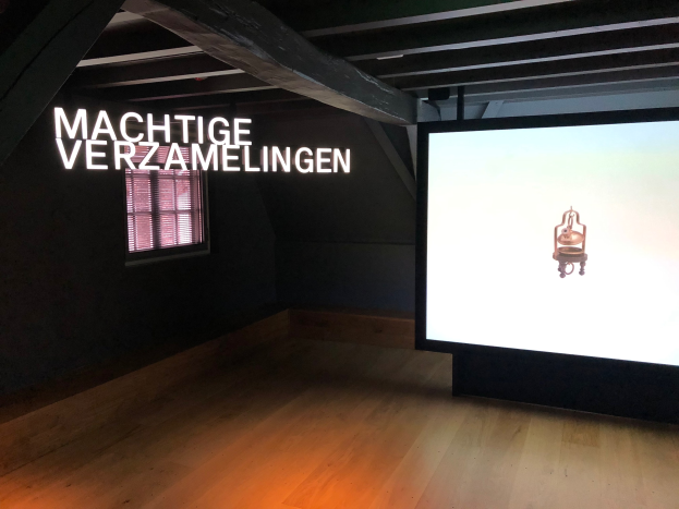 Installationsansicht der Ausstellung "Machtige Verzamelingen" im Museum of Modern Art in Berlin mit einem großen Bildschirm auf der rechten Seite und Text an der Wand dahinter sowie einem Fenster auf der linken Seite.