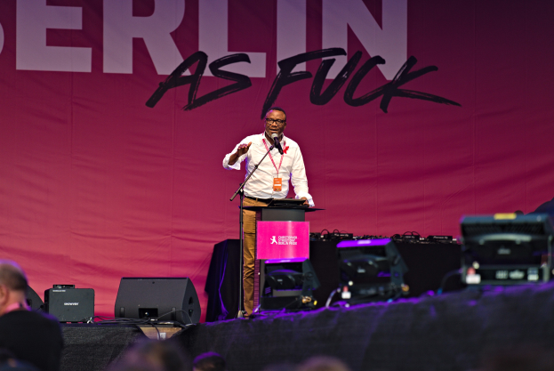 Ein Mann in einem weißen Hemd und braunen Hosen steht auf einer Bühne und hält ein Mikrofon in der Hand, während er eine Menge anspricht. Im Hintergrund ist ein pinkfarbenes Banner mit der Aufschrift "Berlin as Fuck" zu sehen.