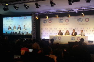 Menschen sitzen auf Stühlen an einem Tisch mit Namensschildern und Mikrofonen auf der Bühne, mit einem Publikum auf Stühlen davor, einem Projektionsschirm und Beleuchtung an Ständern im Hintergrund.