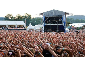 Menge bei einem Musikfestival mit erhobenen Händen, einige halten Handys, vor einer Bühne mit Performern, weißen Zelten und Bäumen unter einem klaren blauen Himmel.