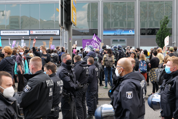 Große Menschenmenge vor einem Gebäude protestierend, einige halten Schilder und tragen Helme, mit einem Pfahl und Schild im Vordergrund und einem Baum im Hintergrund.
