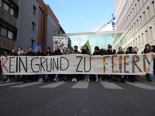 Demonstranten halten ein Transparent mit der Aufschrift "Kein Grund zu Feiern" gegen die deutschen Sparmaßnahmen, mit Gebäuden, einem Schild, einem weiteren Transparent, einem Kran und einem bewölkten Himmel im Hintergrund.