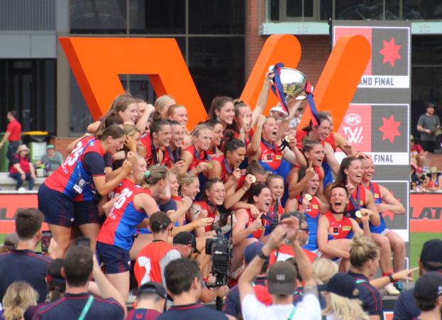 Eine Gruppe von Frauenfußballerinnen in blauen und roten Uniformen feiert mit einem Pokal vor einer jubelnden Menge auf einem Rasenfeld, mit Zuschauern und Sponsorplakaten im Hintergrund.