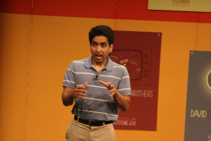 Ein Mann steht auf einer Bühne und spricht, mit einer Wand voller Plakate dahinter.