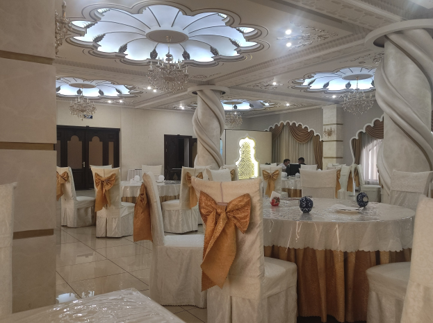 Ein großer Festsaal mit Tischen und Stühlen, die mit Tüchern bedeckt und mit verschiedenen Gegenständen geschmückt sind, mit Türen und Vorhängen an den Wänden und beleuchtet von Deckenleuchten und Kronleuchtern.