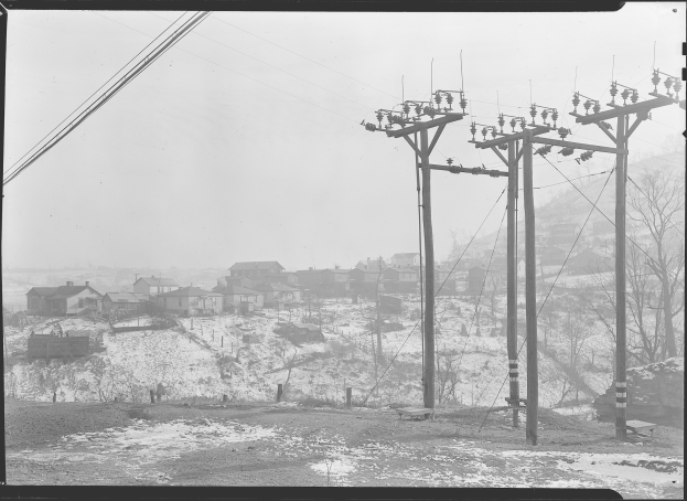 Ein Schwarz-Weiß-Foto einer Stadtlandschaft mit Strommasten, Drähten, Häusern, Bäumen und schneebedeckter Erde.