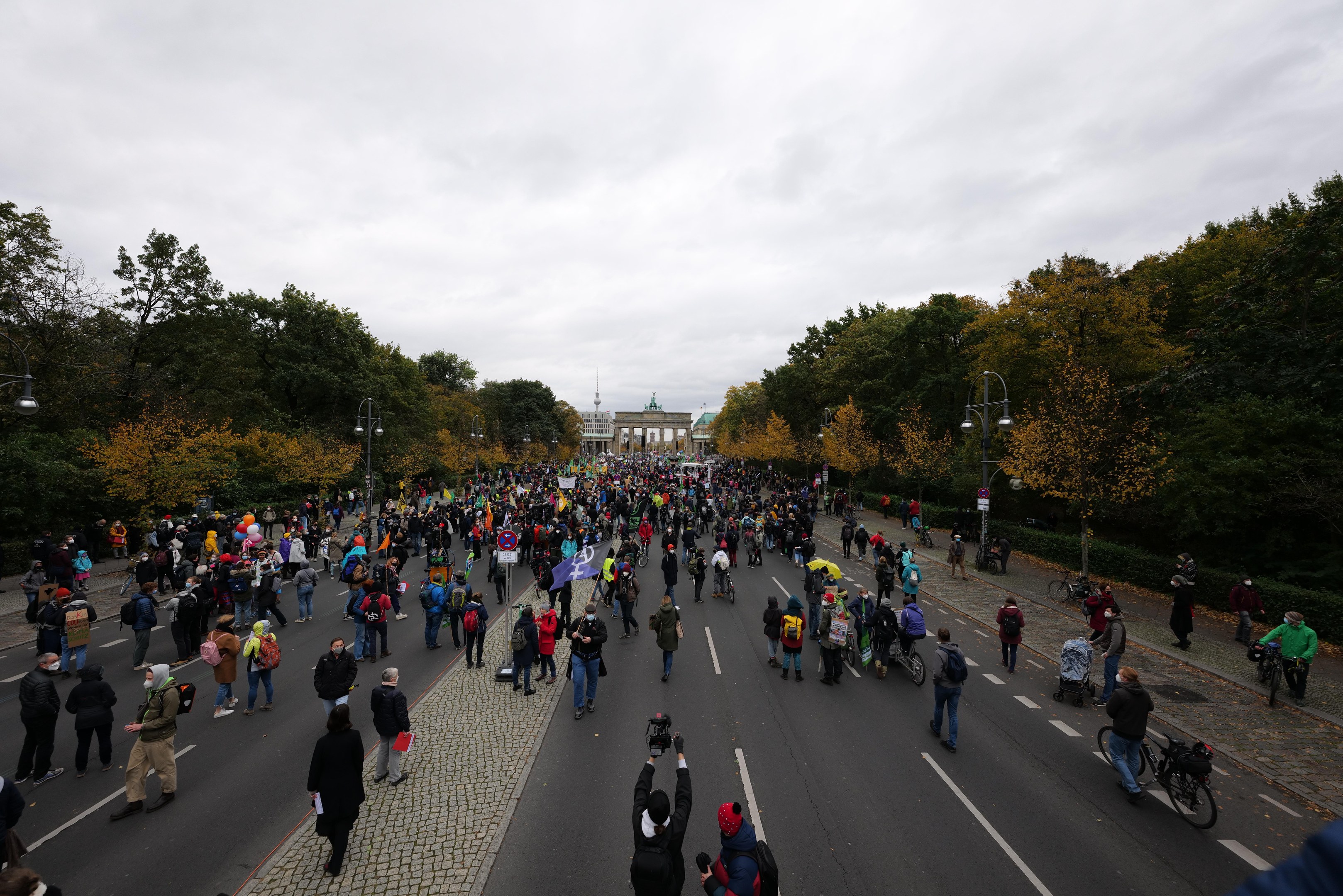Eine große Gruppe von Menschen marschiert eine Baumallee in Berlin entlang, einige halten Kameras, mit einem Gebäude und einem klaren Himmel im Hintergrund.