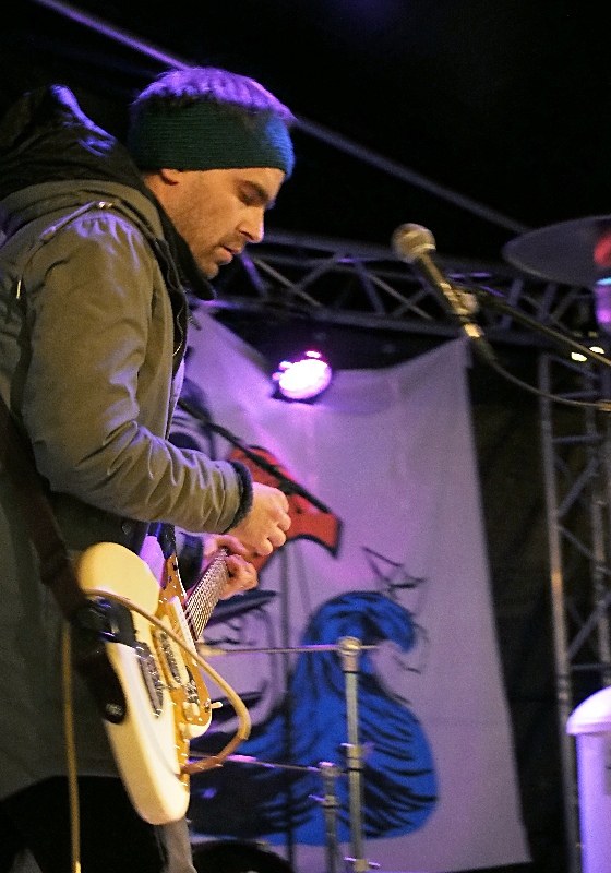 Ein Mann in einer Jacke und einer Mütze spielt eine E-Gitarre auf der Bühne mit einem Mikrofon vor sich, einem Schlagzeug zu seiner Rechten und Scheinwerfern und einem Banner im Hintergrund.