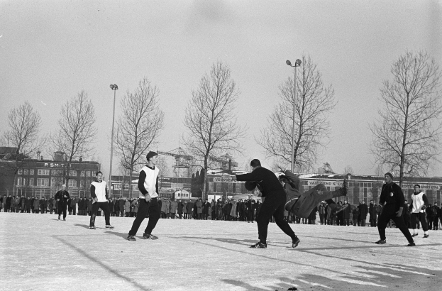Eine Gruppe von Menschen spielt Eis-hockey im Schnee, mit Bäumen, Gebäuden, Laternen und einem klaren Himmel im Hintergrund, dargestellt in Schwarz-Weiß.
