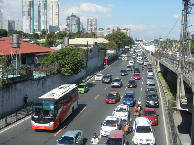 Eine belebte Straße mit Autos, Bussen und Motorradfahrern, die eine Brücke mit elektrischen Mästen und Kabeln überqueren, umgeben von Gebäuden, Bäumen und einem bewölkten Himmel.