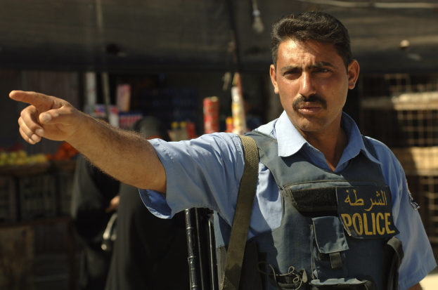 Polizeibeamter in Uniform, der mit einer Waffe vor einem Geschäft steht, mit unscharfen Menschen und Gegenständen im Hintergrund.