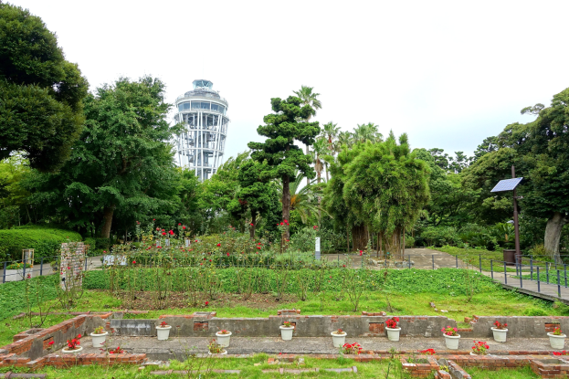 Ein Park mit einem Turm im Hintergrund, umgeben von grünem Gras, Pflanzen, Bäumen, verstreuten Blumentöpfen, einer Straße mit Geländern und einem Pfahl mit einer Tafel, unter einem sichtbaren Himmel.