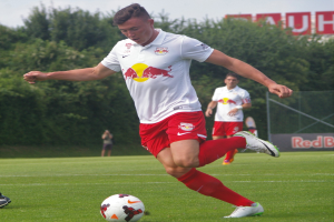 Ein Mann in einem Fußballtrikot schießt einen Ball auf einem grünen Feld mit Bäumen und einem klaren blauen Himmel im Hintergrund, wobei ein Schild mit der Aufschrift "RB Leipzig" dahinter zu sehen ist.