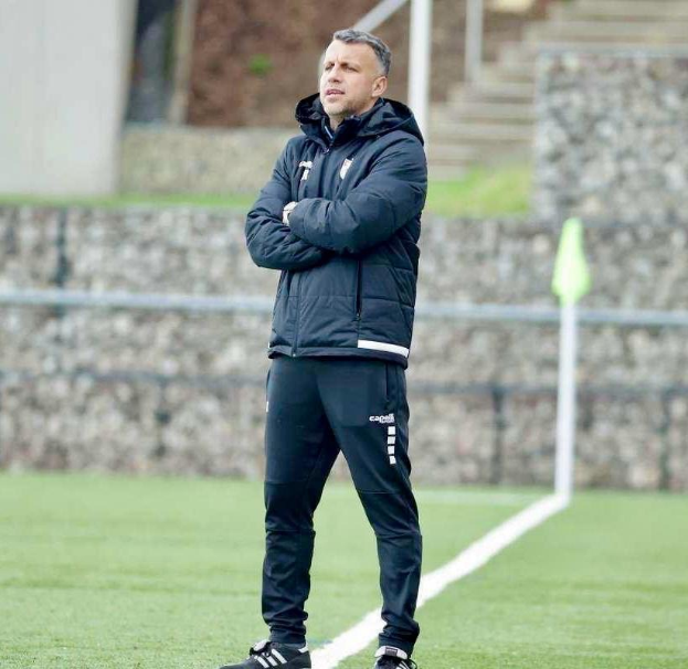 Ein Fussballtrainer, der mit überkreuzten Armen auf einem Feld steht und eine schwarze Jacke und Hose trägt, vor einem Zaun, einer Treppe und einer Wand.