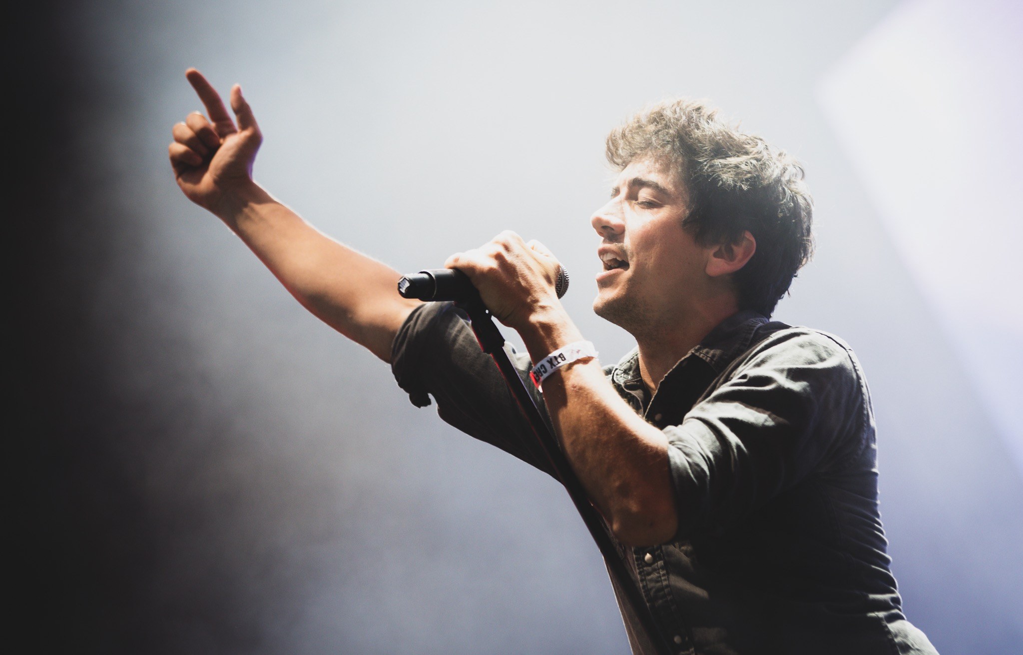 Ein Mann in schwarzem Hemd und Jeans singt leidenschaftlich in ein Mikrofon auf einer hell erleuchteten Bühne, seine Augen sind geschlossen und seine Haare sind unordentlich gestylt, während das Publikum im Hintergrund applaudiert.