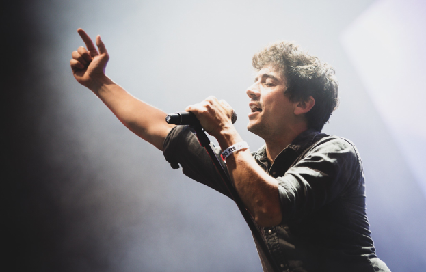 Ein Mann in schwarzem Hemd und Jeans singt leidenschaftlich in ein Mikrofon auf einer hell erleuchteten Bühne, seine Augen sind geschlossen und seine Haare sind unordentlich gestylt, während das Publikum im Hintergrund applaudiert.