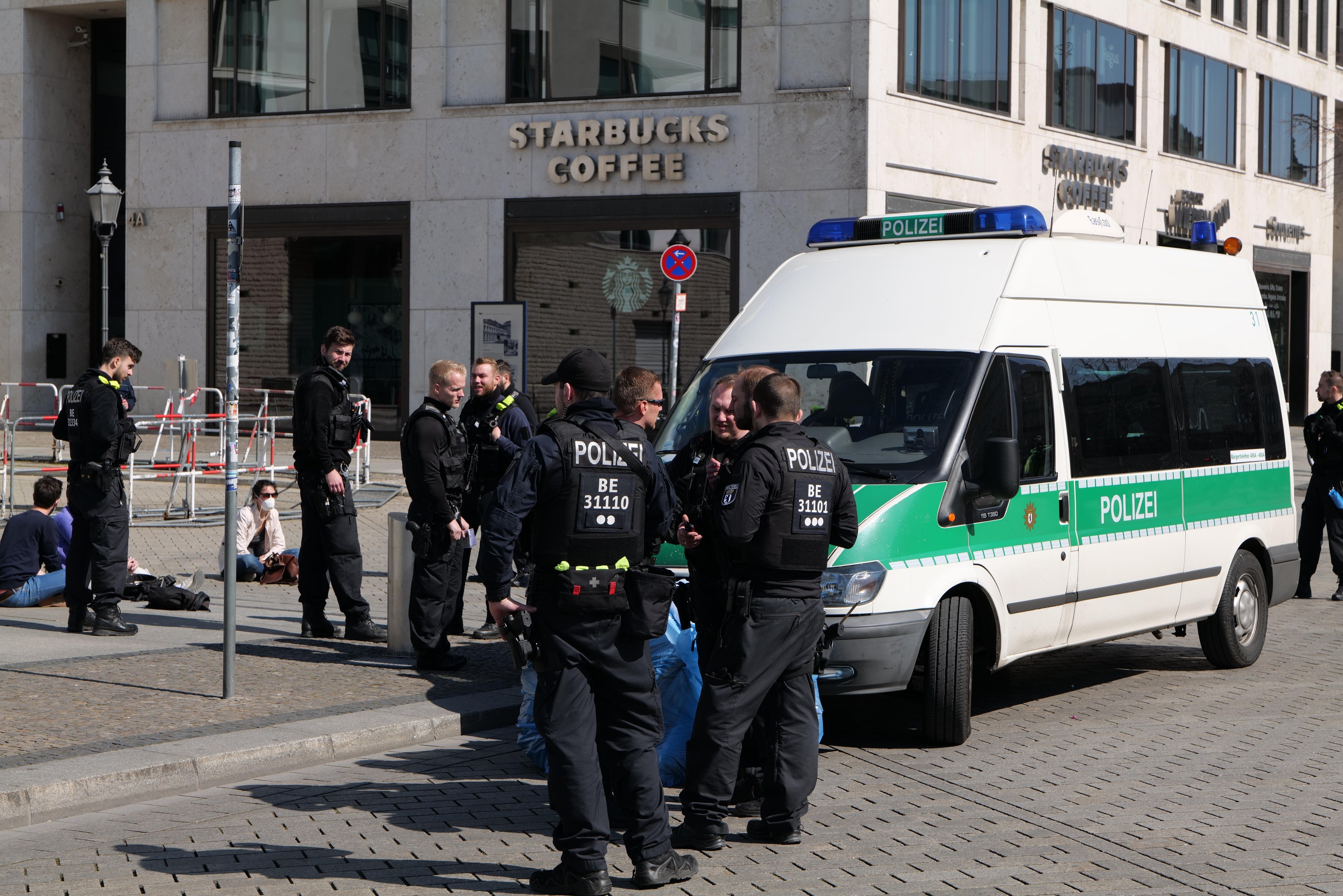 Eine Gruppe von Polizisten steht vor einem Starbucks-Café mit einem Van auf der rechten Seite und einigen Menschen auf der linken Seite; im Hintergrund sind ein Gebäude, ein Schild, ein Lichtmast und ein Zaun zu sehen.