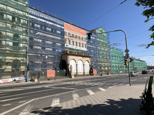 Großer mehrstöckiger Bau im Bau in Berlin mit zahlreichen Fenstern, umgeben von Straßeninfrastruktur einschließlich Masten, Laternen, Signalen, Fahrzeugen, Fußgängern, Werbetafeln, Bäumen und einem bewölkten Himmel.