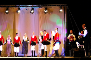 Eine Bühnenperformance mit Menschen auf der Bühne, ein Vorhang im Hintergrund, Scheinwerfer oben, Musiker rechts und ein weißer Stuhl in der rechten unteren Ecke.