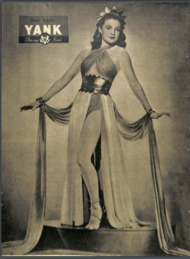 Ein Schwarz-Weiß-Foto der Schauspielerin Joan Leslie in einem langen, hochgeschlossenen Kleid mit langen Ärmeln, die selbstbewusst auf einer Bühne steht, die Augen geschlossen, mit 1940er-Jahre-Stil-Haare und Make-up.