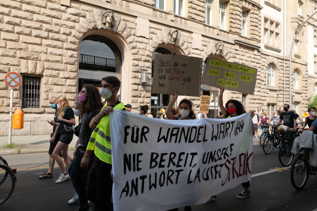 Eine Gruppe von Menschen marschiert bei einer Demonstration gegen die Anti-Kriegs-Bewegung in Berlin, einige halten Schilder und andere fahren Fahrräder, mit einem Gebäude im Hintergrund, das Fenster, Bögen, Säulen und Skulpturen aufweist.